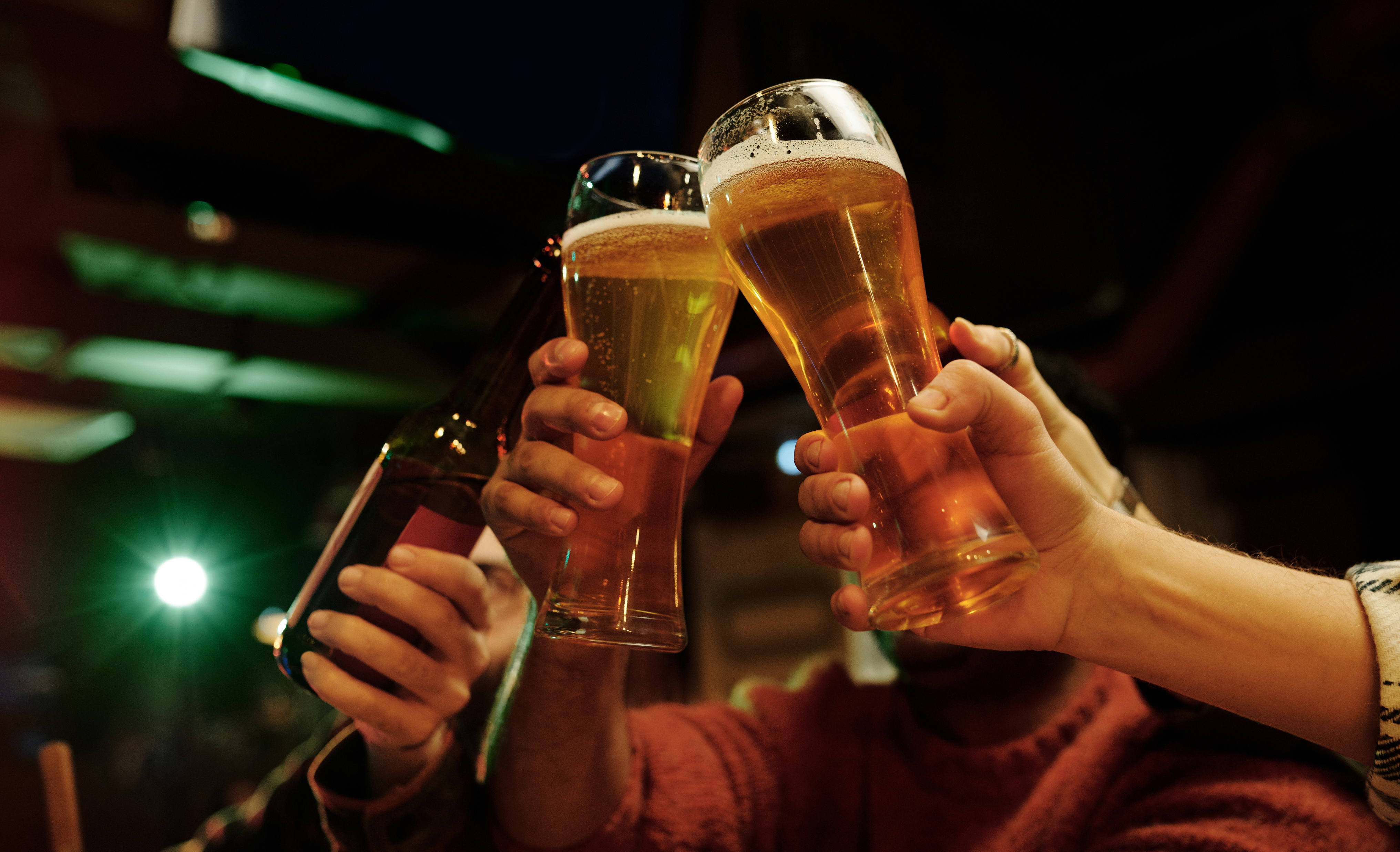 três pessoas brindando com dois copos de cervejas e uma long neck em um pub com luz esverdeada