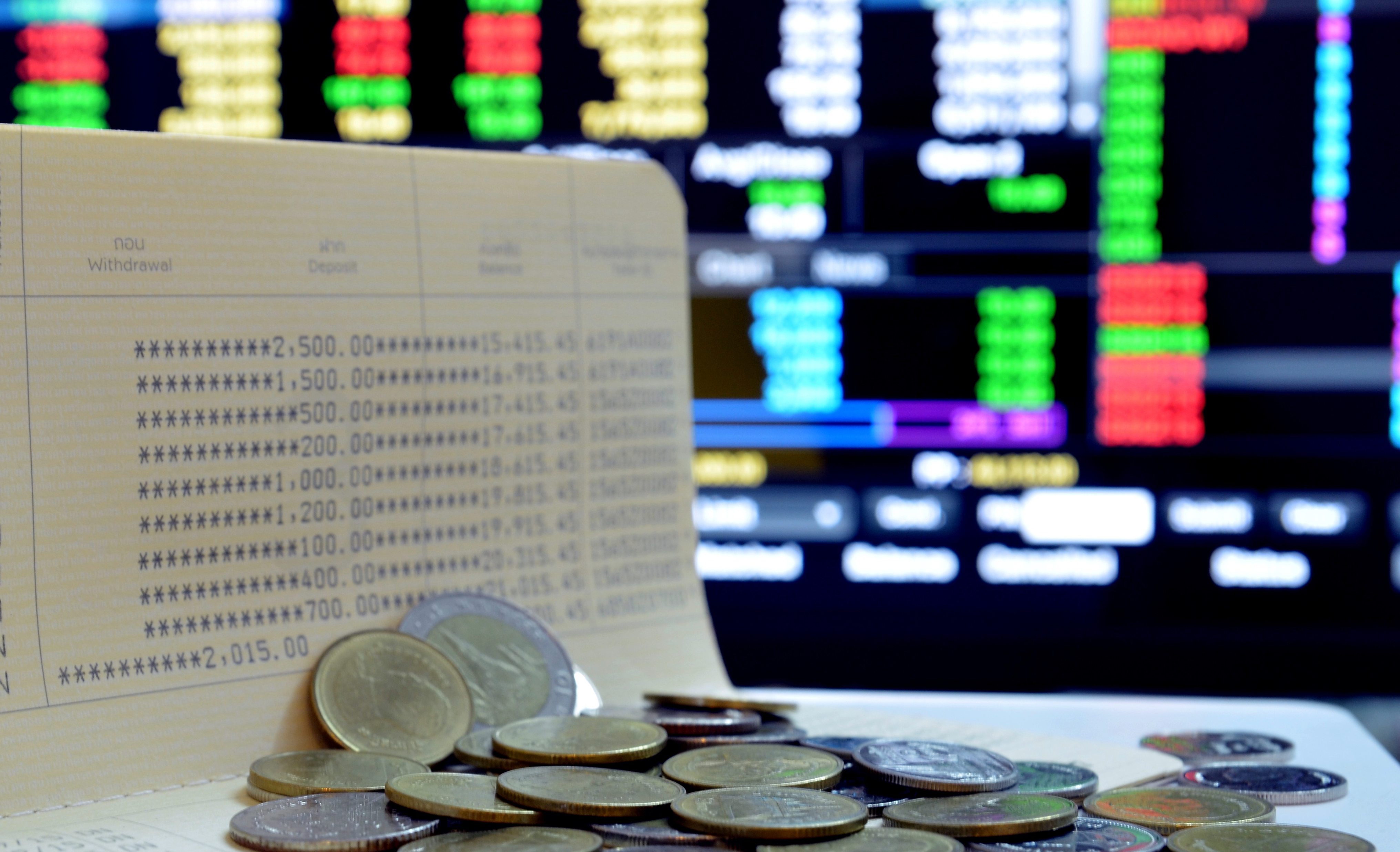 moedas espalhadas sobre um papel onde gastos estão escritos, ao fundo um painel de investimentos, simulando a bolsa de valores